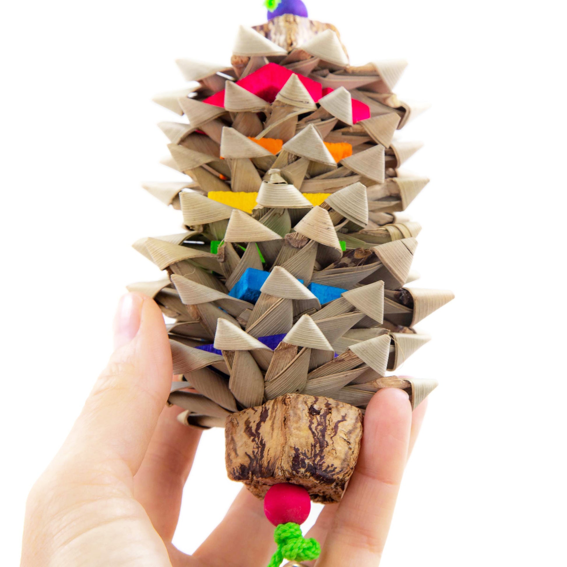 Colorful bird toy with rainbow order thin wood chipper blocks alternating with woven palm flowers. Top and bottom of the toy ends with a mahogany pod slice and a wooden bead. Shown in a hand for scale.